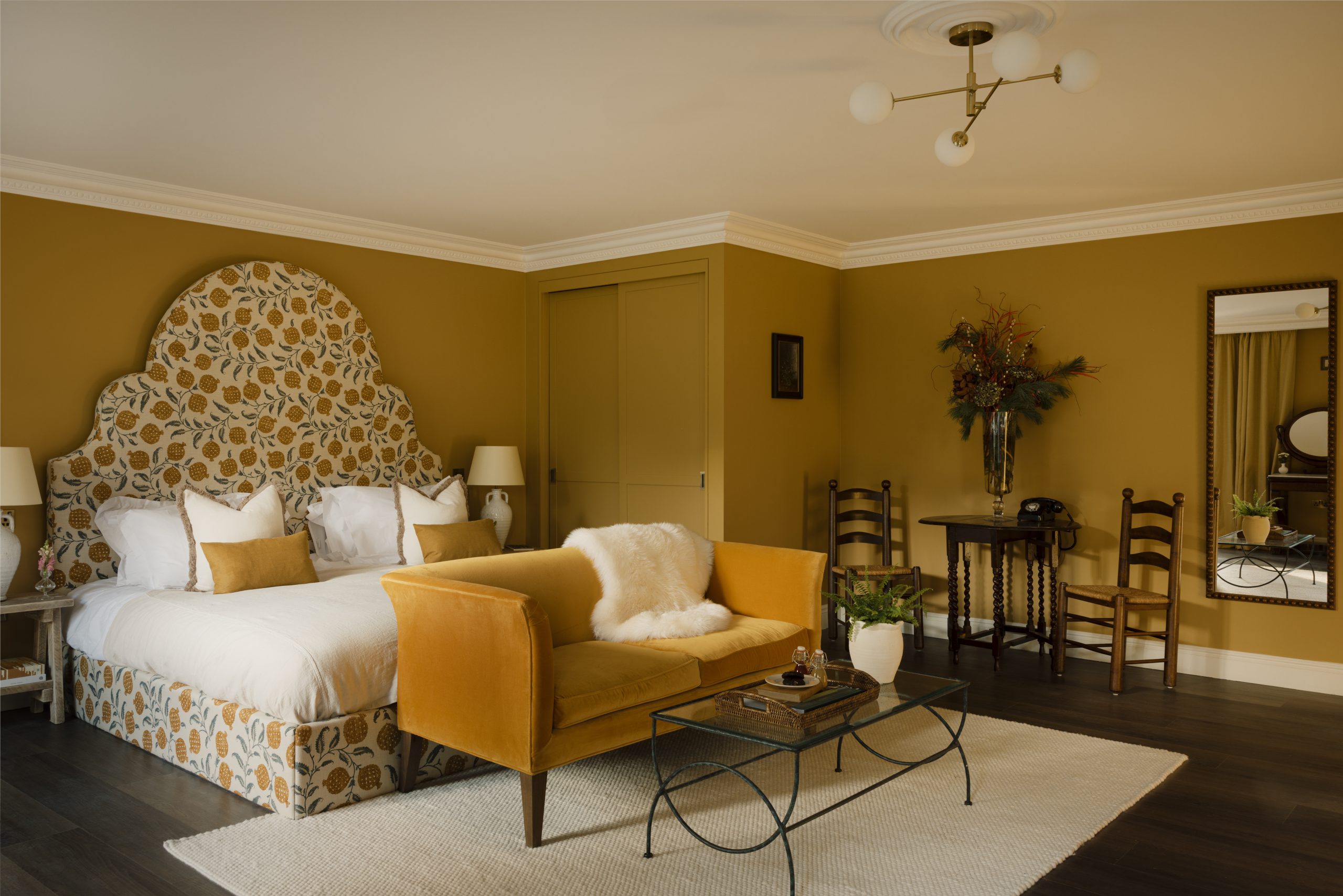 An ochre-tinted, country-style hotel bedroom features spherical, multi-part chandeliers in brass and glass, a velvet couch in the same color of the walls, a huge headboard decorated with floral patterns, wooden furniture, and a wrought iron coffee table.