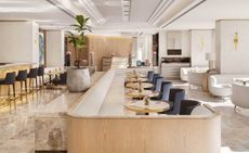 Interior view of a restaurant and bar at The One restaurant, Spain featuring light coloured walls, tile flooring, an inverted ceiling design, a green plant in a pot, pink flowers in vases, round tables, black and gold chairs, white sofas and wooden and off-white coloured seating