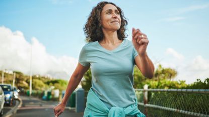 Woman walking outdoors along a sunny road with trees in activewear with sweater tied around her waist, after wondering is walking enough exercise