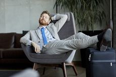 A business man reclines in a chair while resting his feet on his luggage.