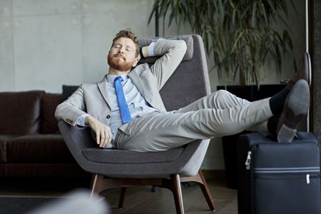 A business man reclines in a chair while resting his feet on his luggage.