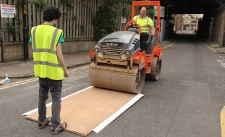 Two men in hi-vis clothing with one driving a rolling machine and the other holding the printing press in place by standing on it. They are in the middle of a road