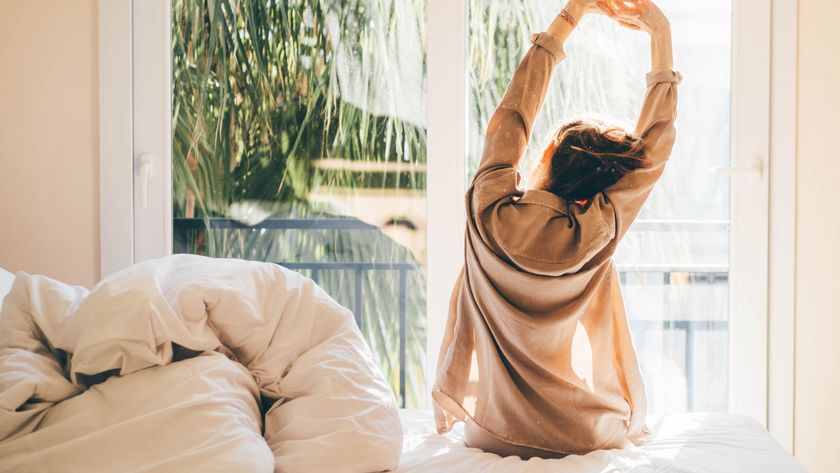 A woman sits on the edge of her bed facing the window, stretching in the morning light shortly after waking up.