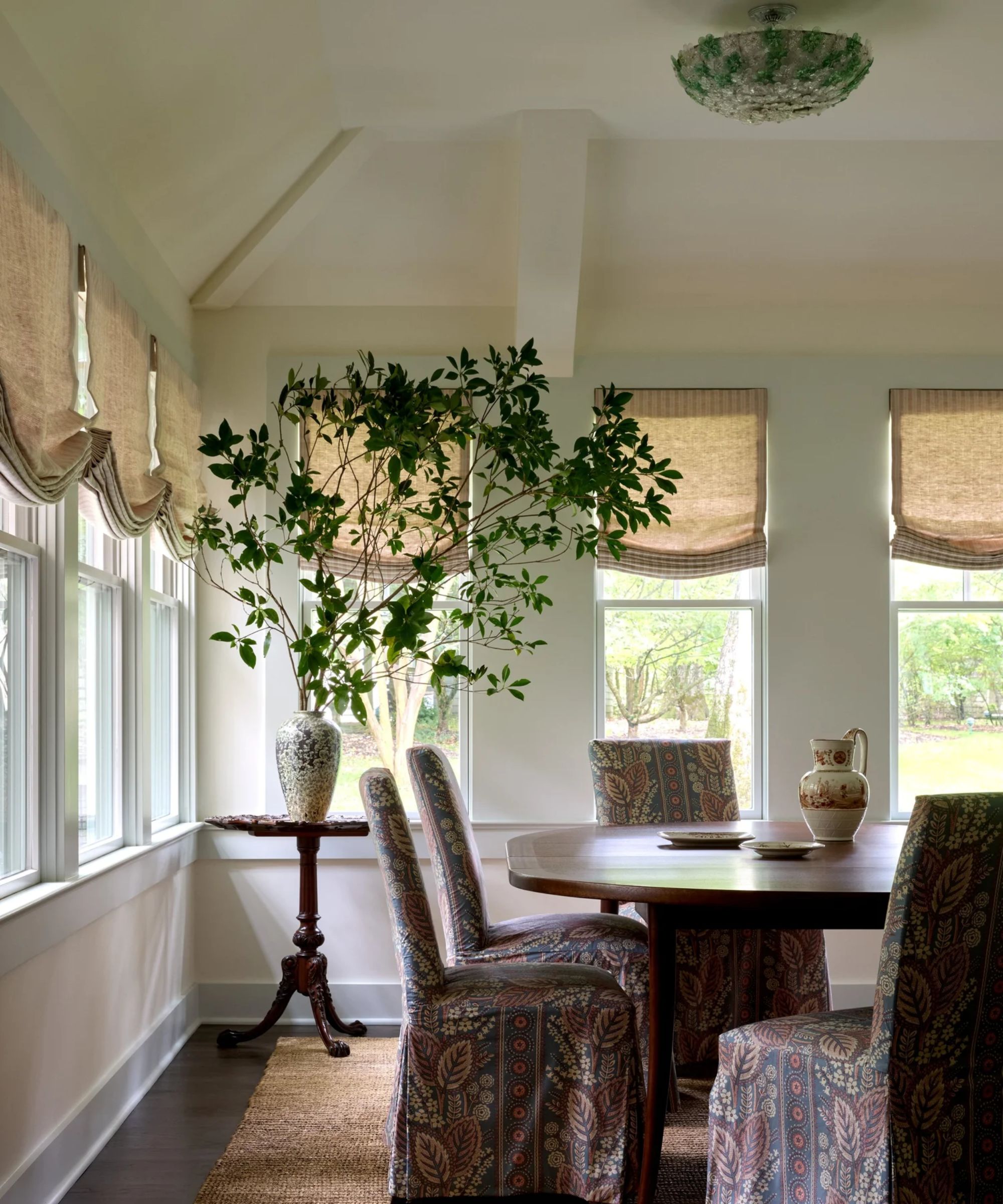 A small sunroom with a round dining table, upholstered dining chairs, linen Roman blinds, a natural jute rug and a large leafy arrangement beside wraparound windows.