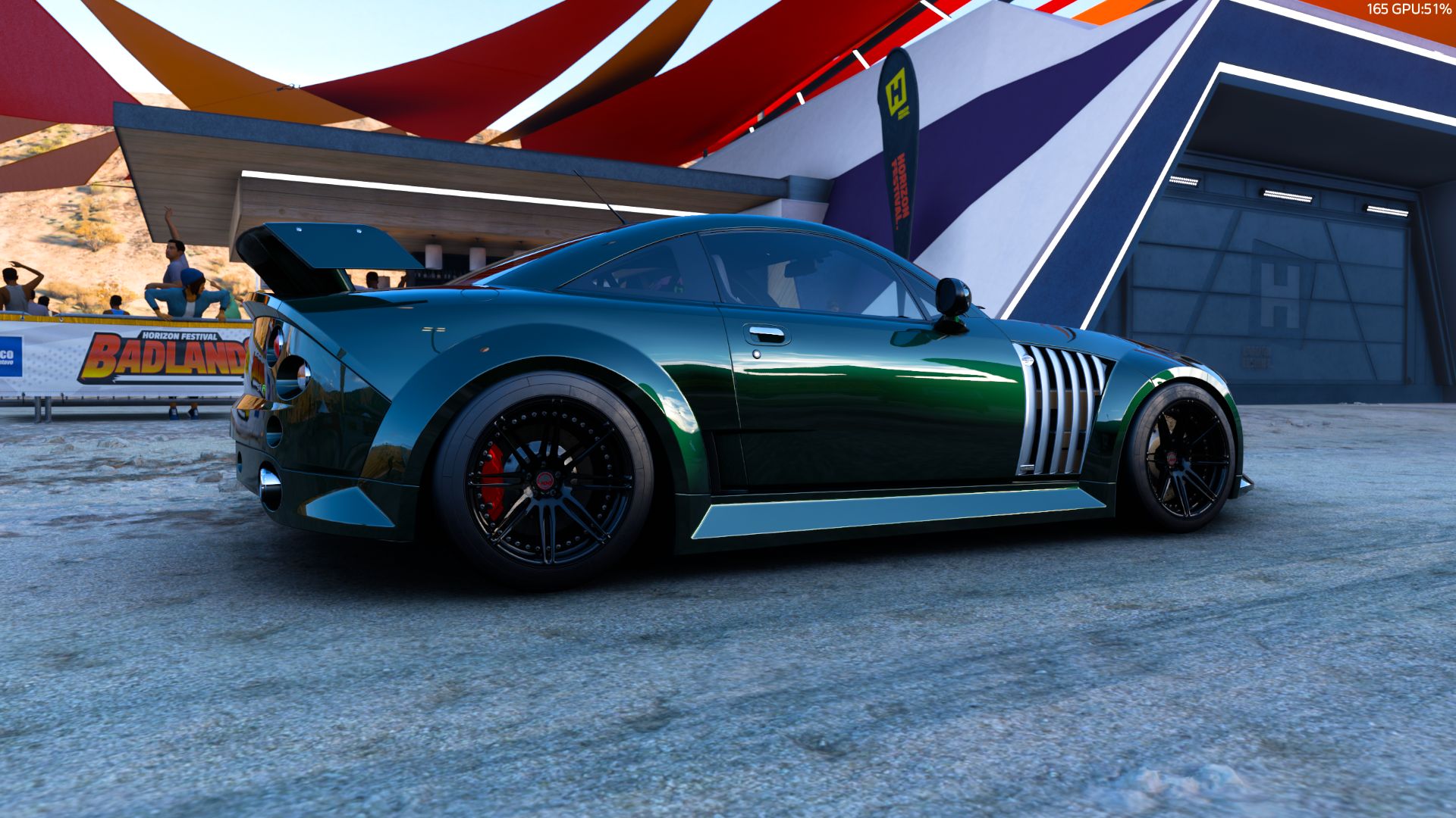 A sleek, dark green MG SV sports car with a large rear spoiler is parked on a concrete surface in Forza Horizon 5. Colorful banners and a modern building are in the background.