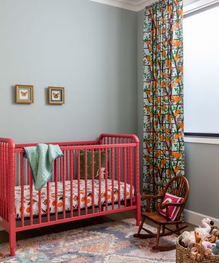nursery with red painted crib, blue painted walls, floral curtains, wooden chair and woven wicker storage basket