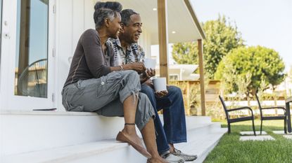 A relaxed, happy older couple sit on their porch steps with coffee.