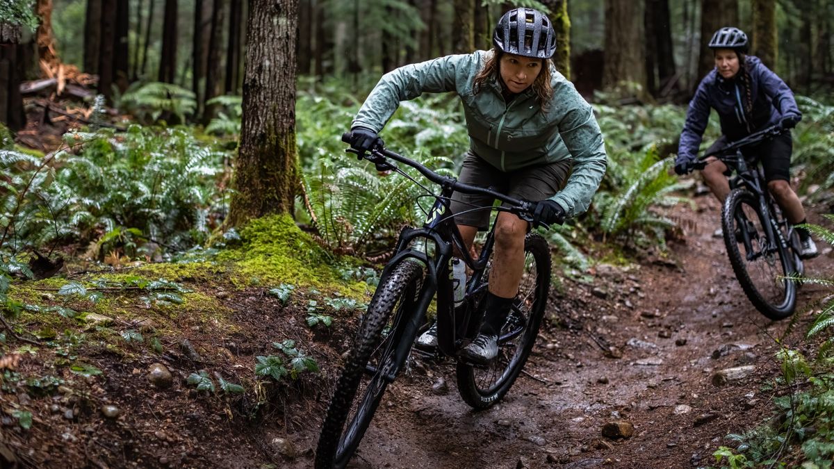 Two women of colour are riding Liv Embolden full-suspension trail bikes through the woods, it&#039;s wet and muddy, and they&#039;re wearing trail jackets and shorts, and have muddy legs