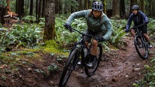 Two women of colour are riding Liv Embolden full-suspension trail bikes through the woods, it's wet and muddy, and they're wearing trail jackets and shorts, and have muddy legs