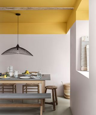 Dining room with off white painted walls and bright yellow painted ceiling, light wood dining table with gray tabletop, matching bench, wooden stools, woven baskets on floor, textured black pendant light