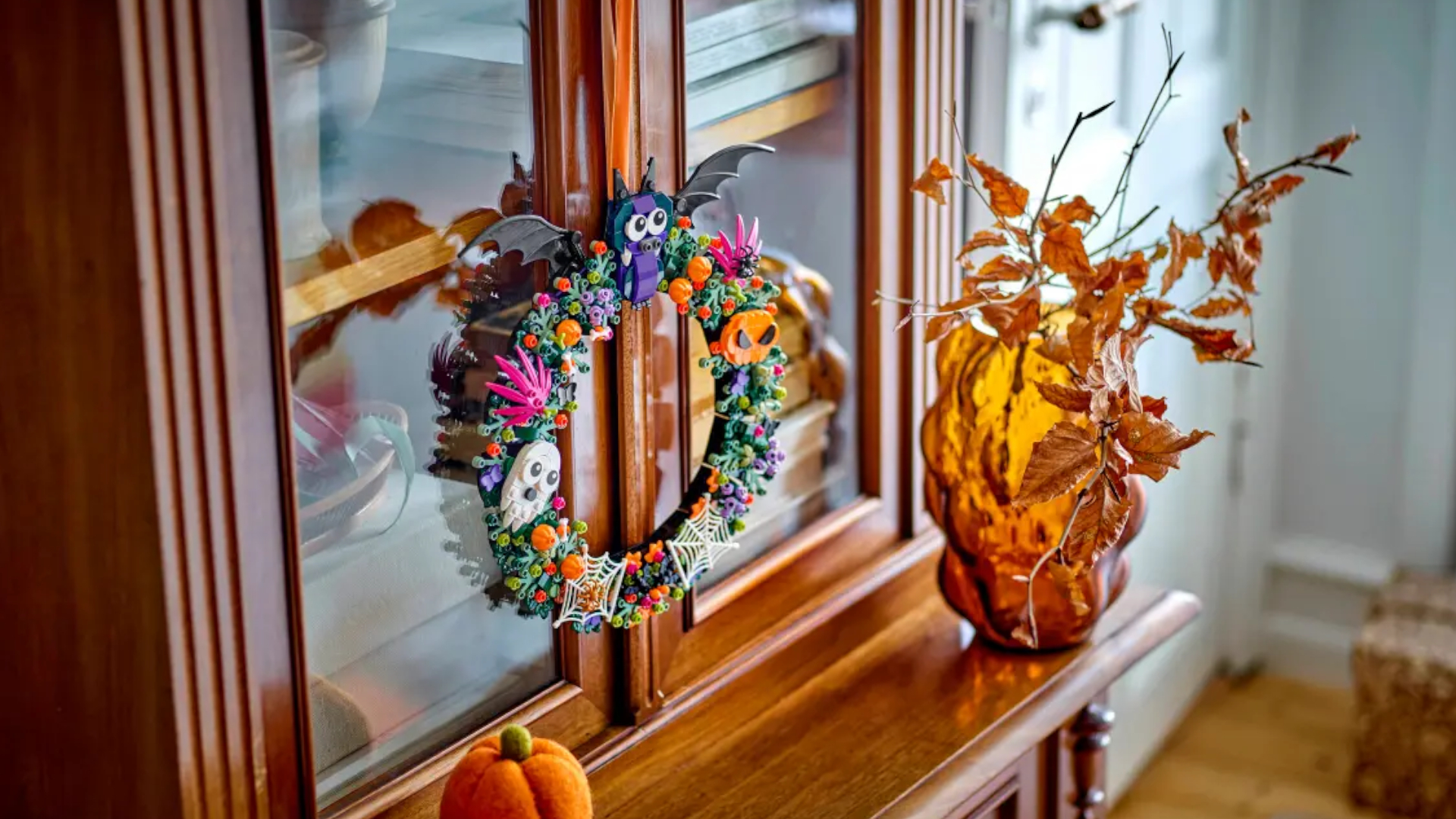 Lego Halloween Wreath hanging on a cabinet, with a plush pumpkin and orange vase nearby
