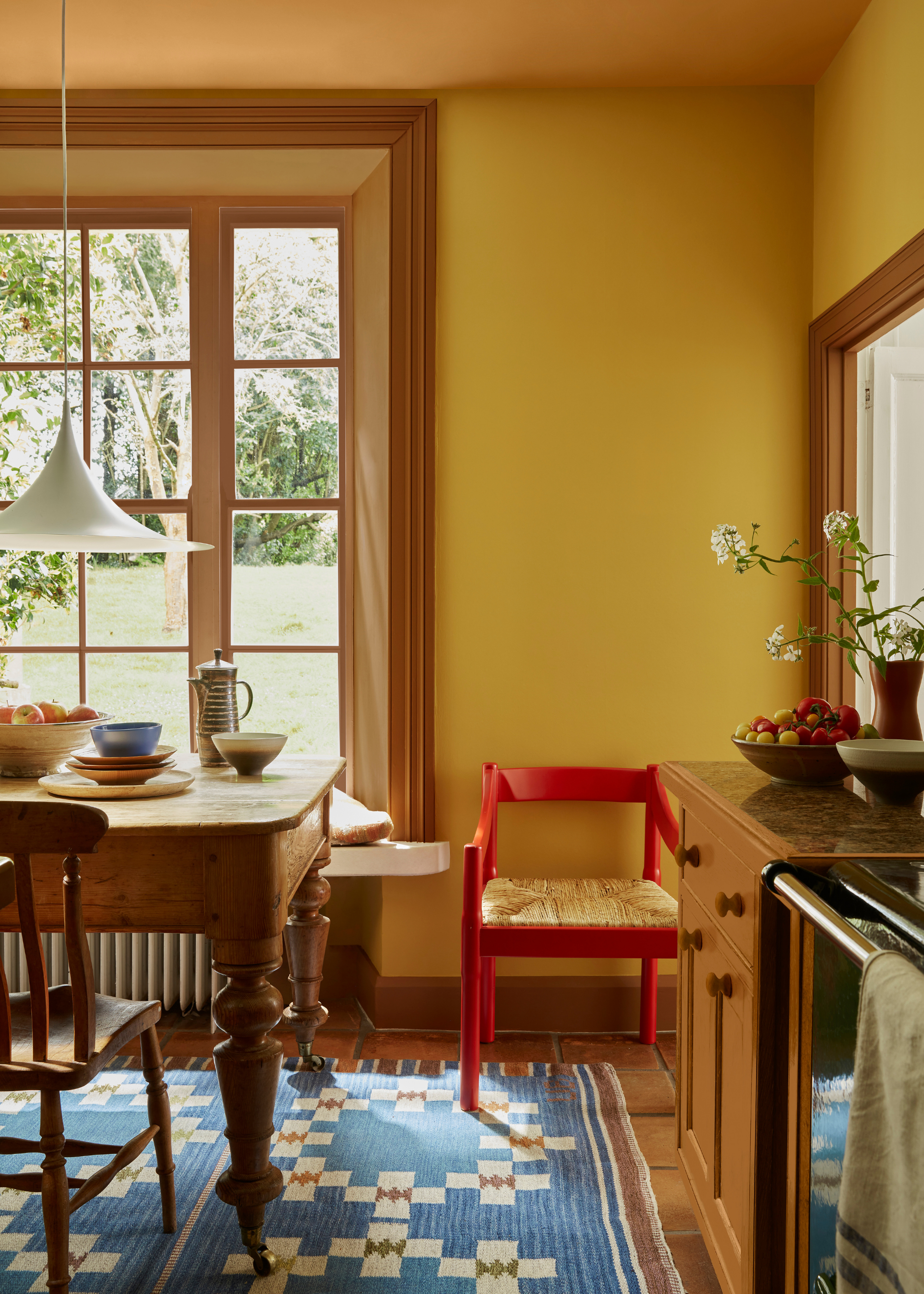 kitchen and dining room with bright yellow wall, red armchair, patterned blue rug, timber-framed windows, a timber dining table and low hanging white pendant