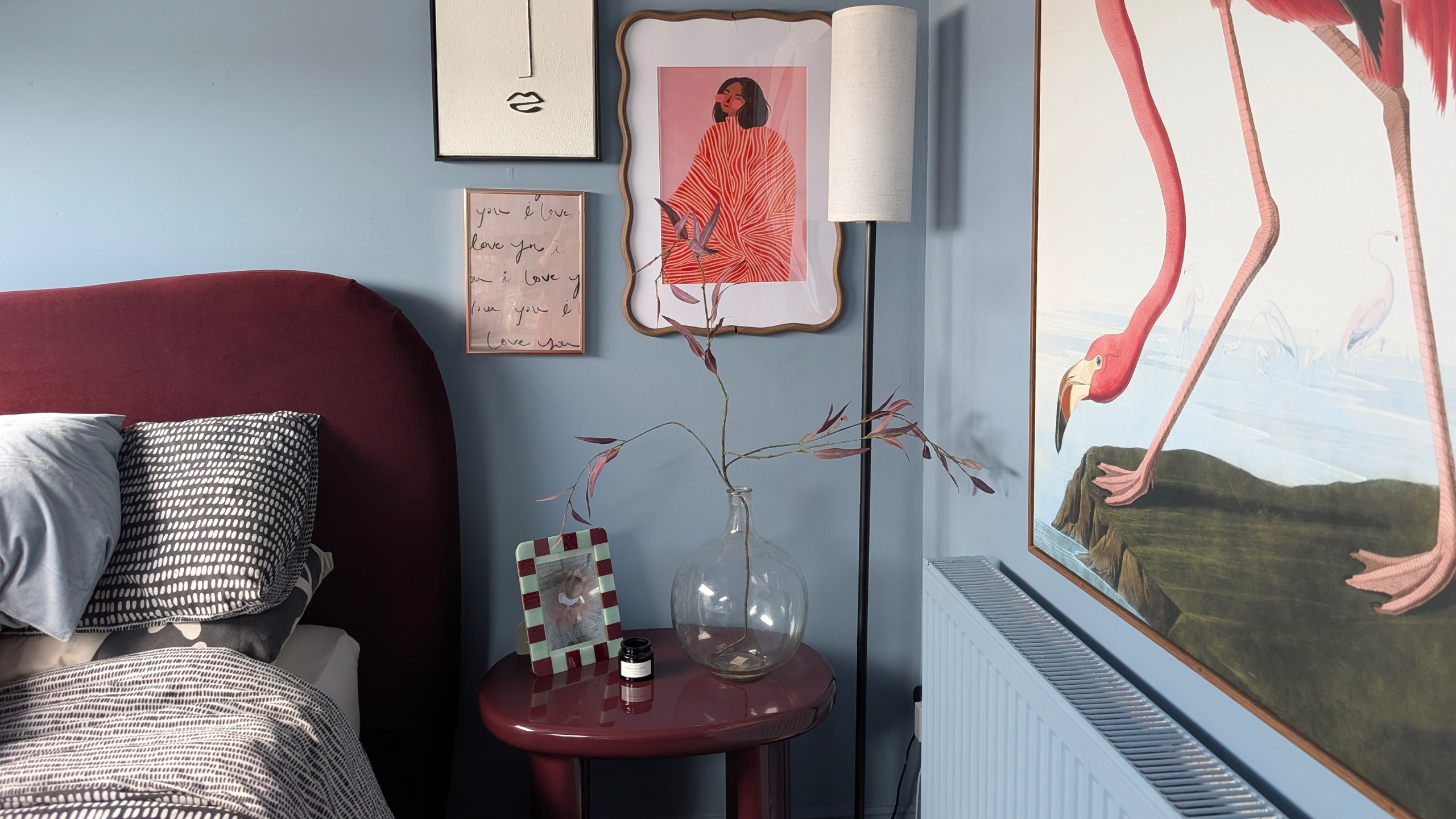 Blue bedroom with burgundy bed and bedside table, trio of framed prints on wall and large painting of flamingo above radiator