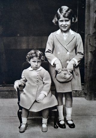 A black and white picture of Princess Margaret sitting in a chair wearing a dress coat and Princess Elizabeth standing next to her and holding a purse