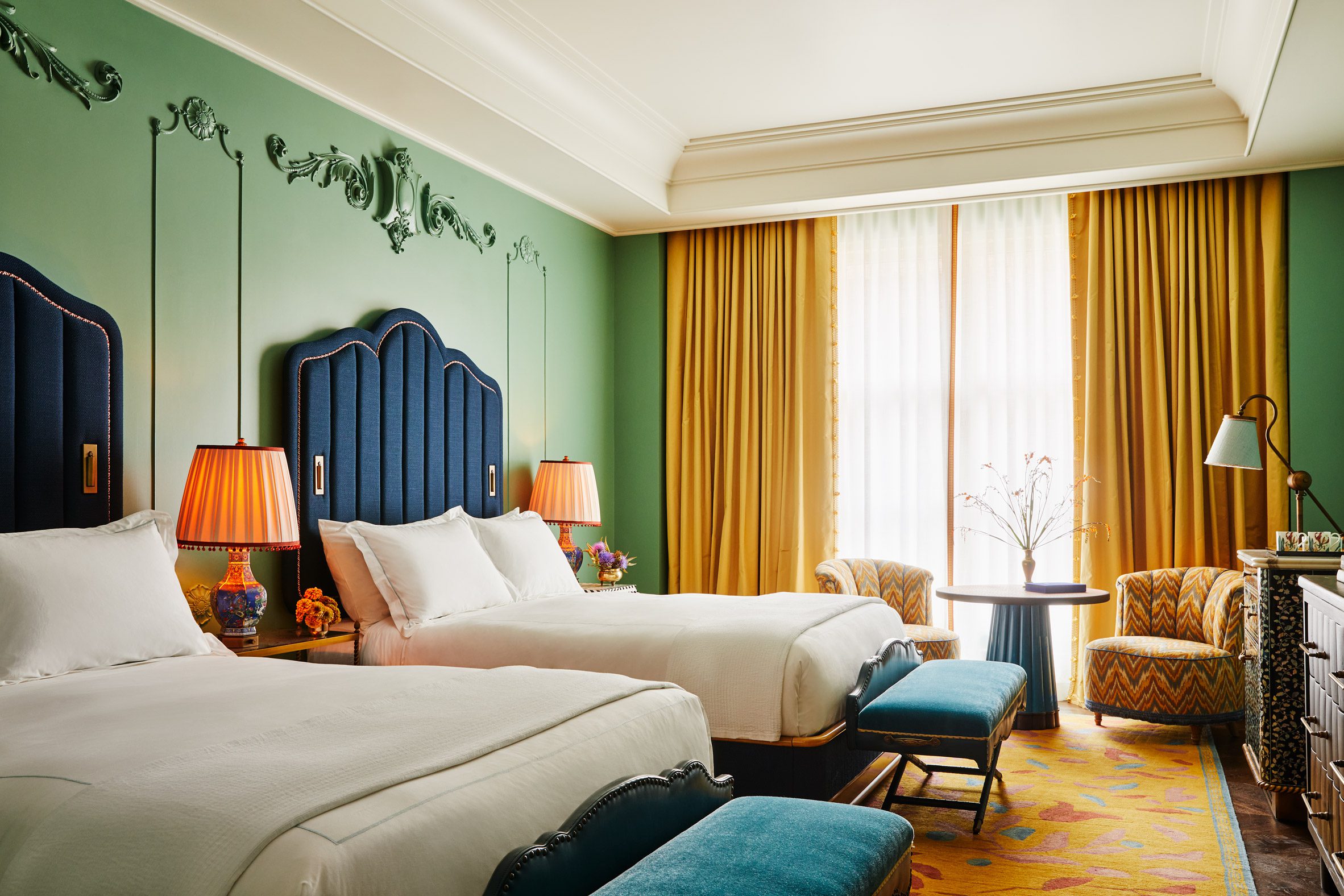 A whimsically decorated, maximalist hotel room is dressed in plush tile, ochre, blue, and terracotta textiles, grand chandeliers and table lamps, and refined upholstery with a nostalgic, retro twist.