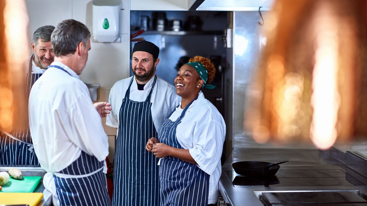 Four chefs stand around in a kitchen smiling and talking.