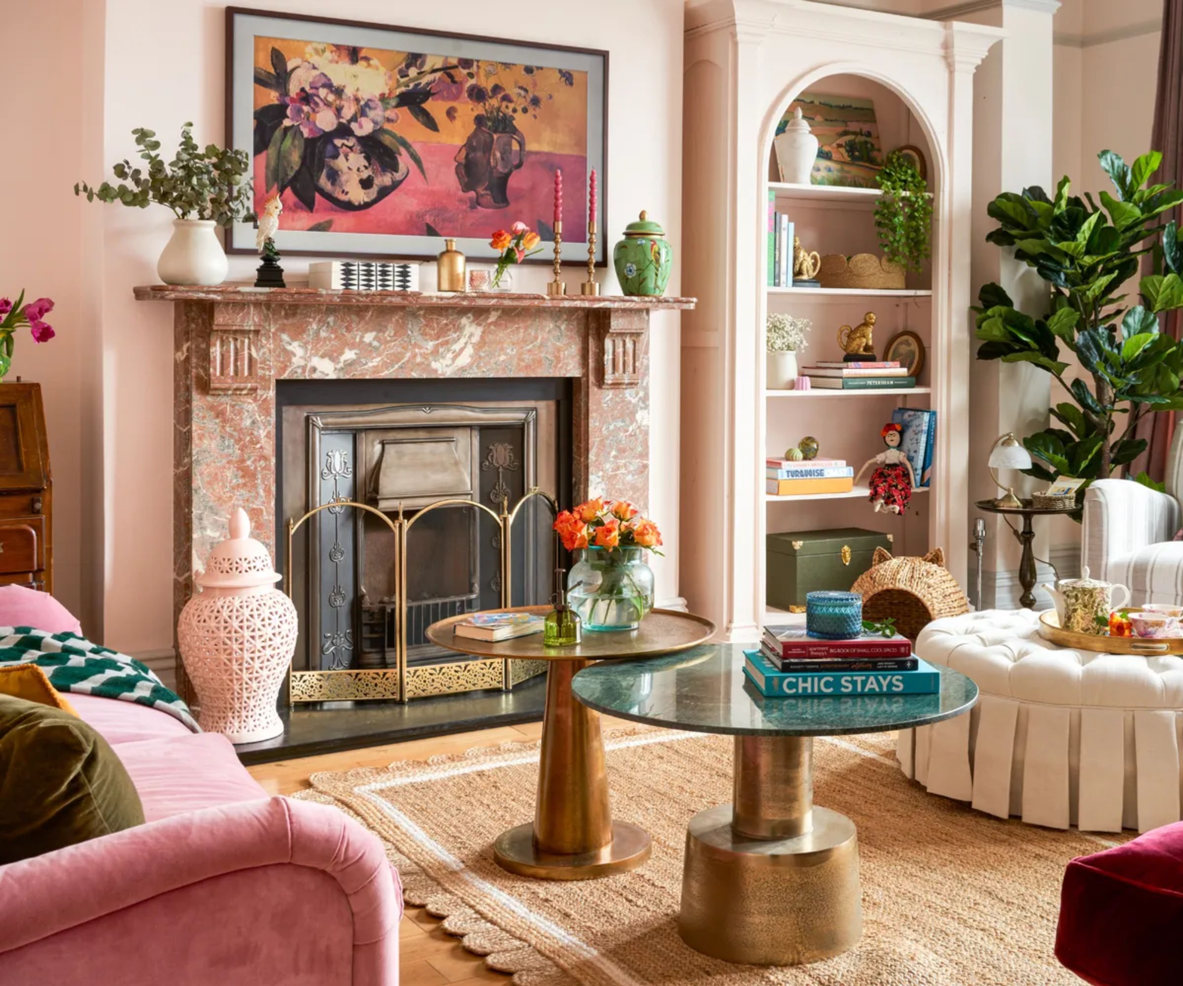 A red marble fireplace with a brass guard sits beneath a colourful floral painting, surrounded by eclectic furniture and decor.