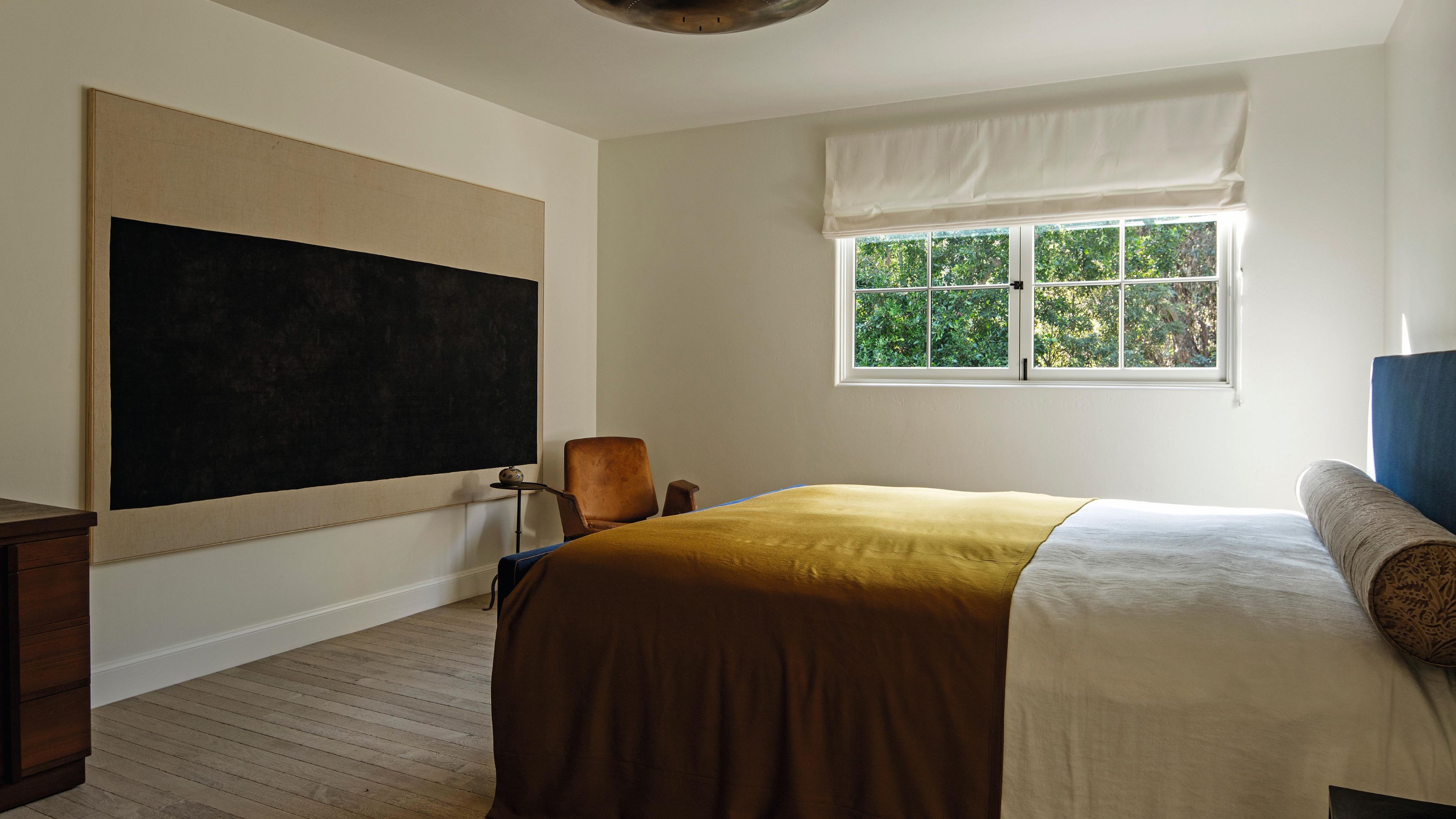 a large neutral bedroom with abstract art and a blue headboard with a mustard throw blanket on the bed