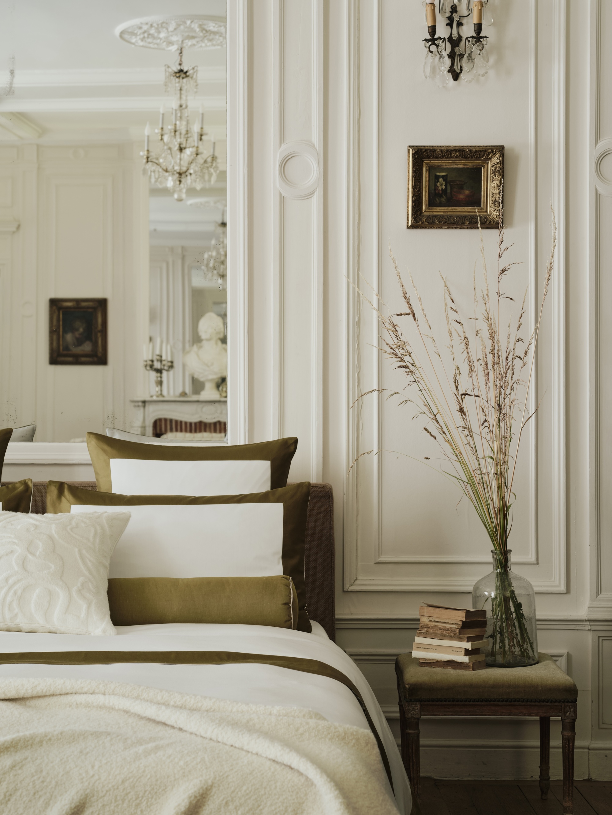 Image of the corner of a luxurious bed against a white wall with intricate molding. The bedding is white with green borders and matching bolster pillows and throw pillows. There is a side table next to the bed with books and a vase.