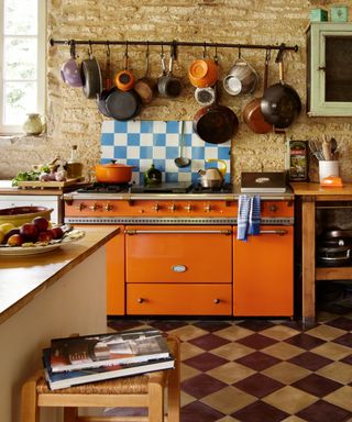 Rustic kitchen with tiled flooring, exposed brick walls, bright orange cooker, pots and pans on rack hanging from wall, kitchen island, blue and white square tiled splashback
