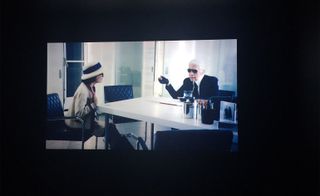 Still of a film against a dark background featuring Karl Lagerfeld sitting at a white desk and a fictional Coco Chanel sitting opposite in a black, diamond-patterned chair