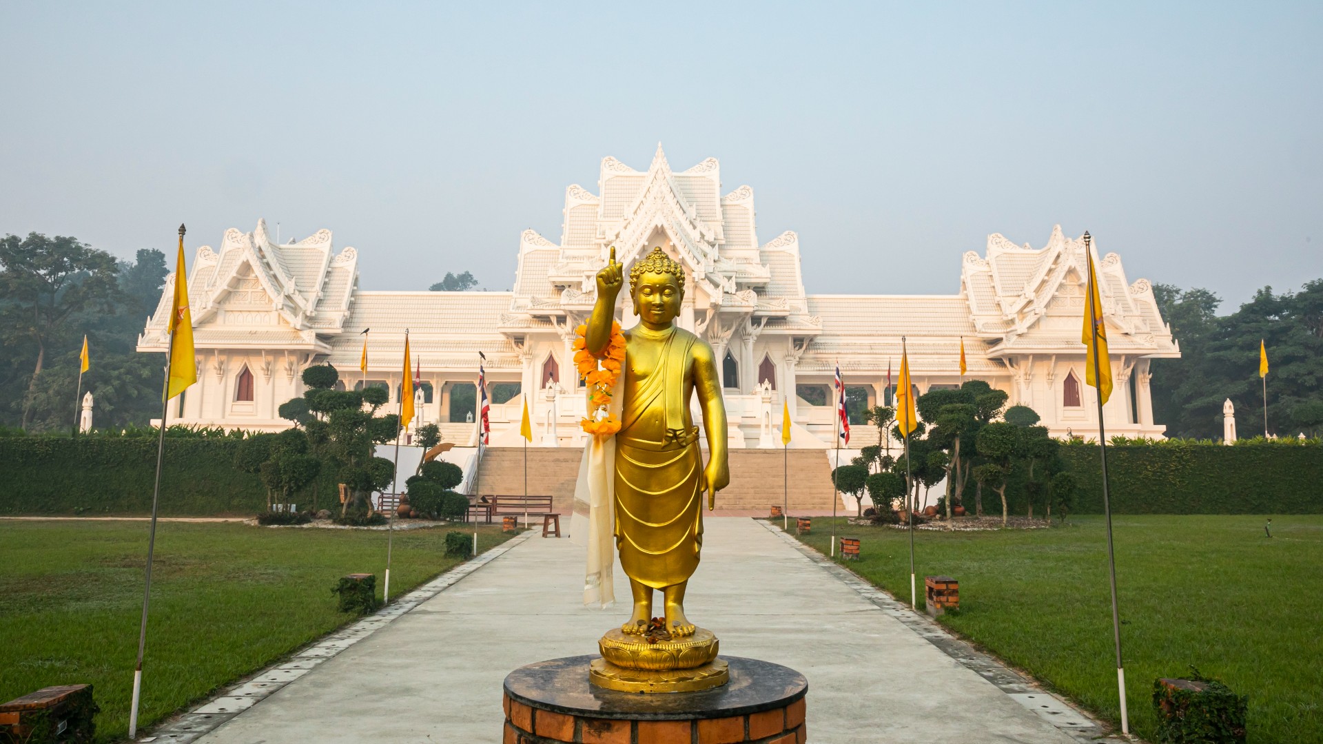 Here is the Royal Thai Monastery in Lumbini, Nepal. Front and center there is a standing golden Buddha statue, with one arm raised pointing upwards. Behind the statue is a white/grey path lined with yellow flags that leads to the white wat-style monastery. It is surrounded by a tall green hedge. The sky is a clear, light blue.