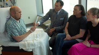 a man in a hospital gown holds up a mirror to see his new facial transplant, while his doctor, dressed in a suit, and his wife and daughter sit and watch nearby