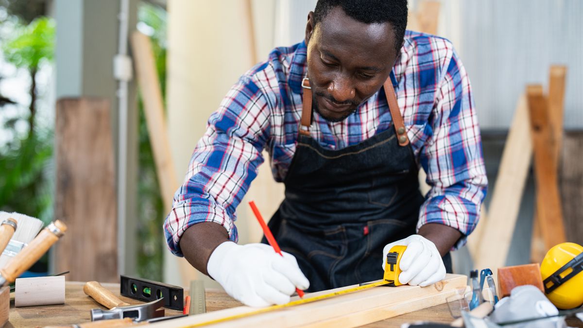 black man using tape measure on piece of wood with tools around him