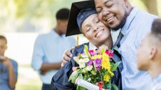 Man gives woman a bouquet of flowers on her graduation day. He is hugging her, while she is holding the flowers and a diploma