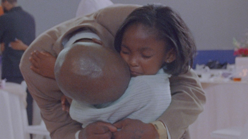 a bald man in a suit leans over and hugs his daughter, who has her eyes closed
