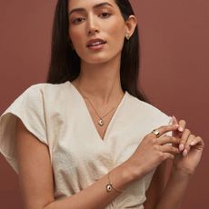A model wearing a white outfit stands infront of a terracotta coloured backdrop wearing layers of stunning Clogau jewellery