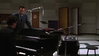 Tom Hanks stands in a music recording studio, leaning on a black piano, in That Thing You Do!