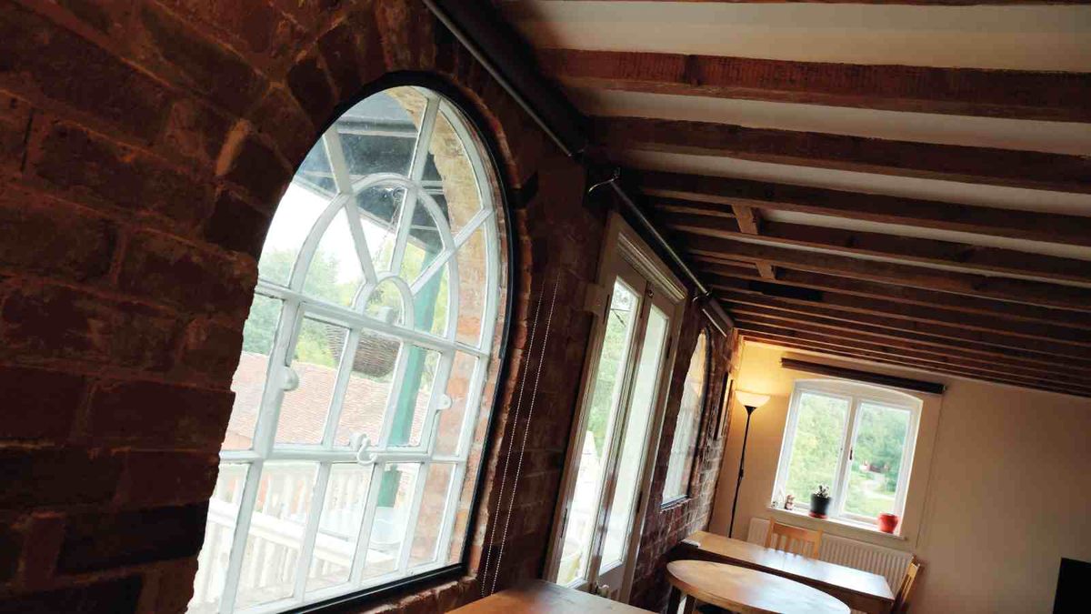 A living room, shot at an angle. The room features a large arched window, exposed brick walls and exposed ceiling beams.