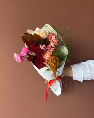 A woman wearing a longsleeved white shirt hold a wrapped bouquet of flowers featuring red roses and a pink orchid and peonies