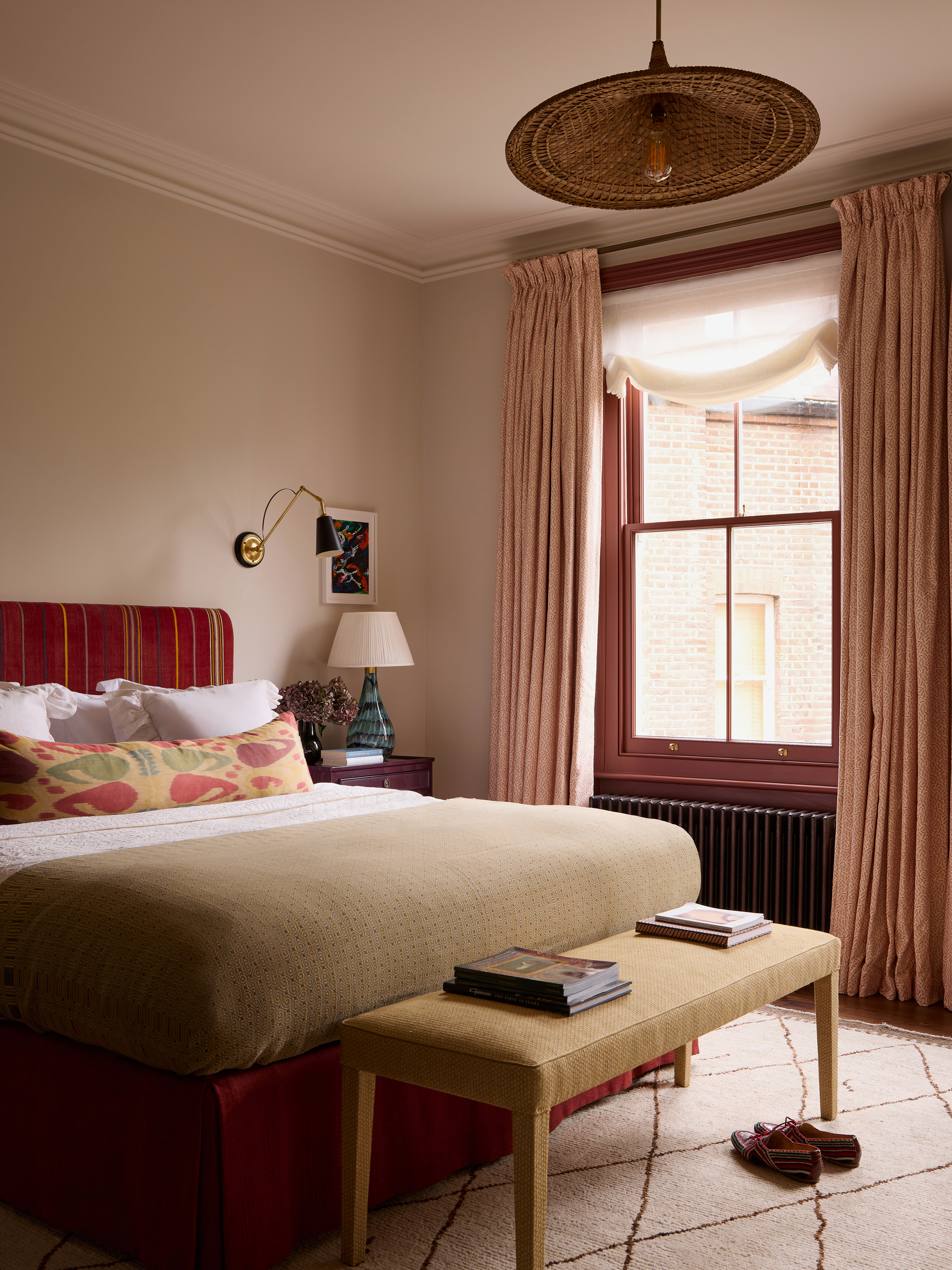 Image of a bedroom with beige walls and a dark red window frame with pink curtains. The bed is red with white and beige bedding.