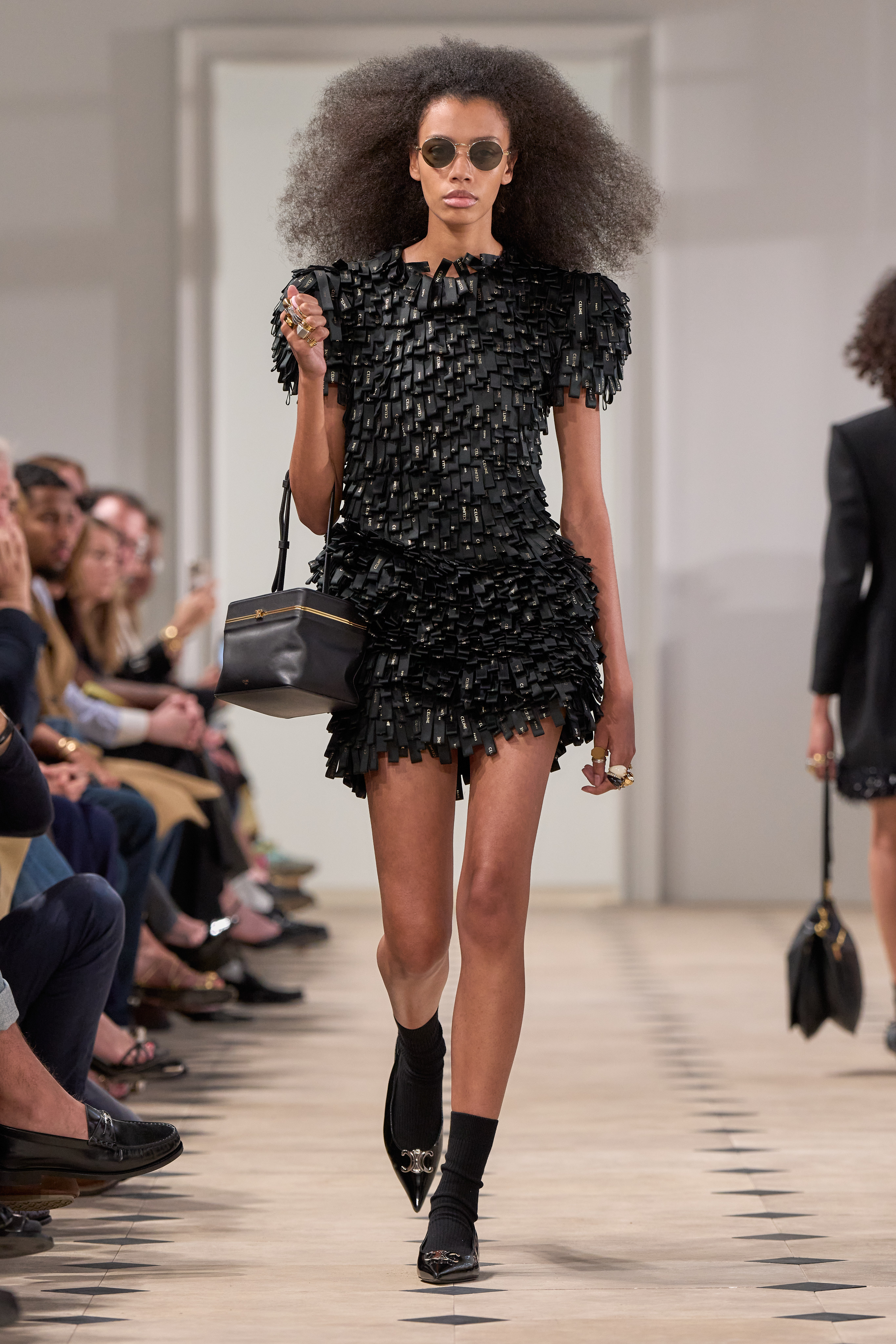 a model walks the Celine runway wearing a dress comprised of several hundred celine labels