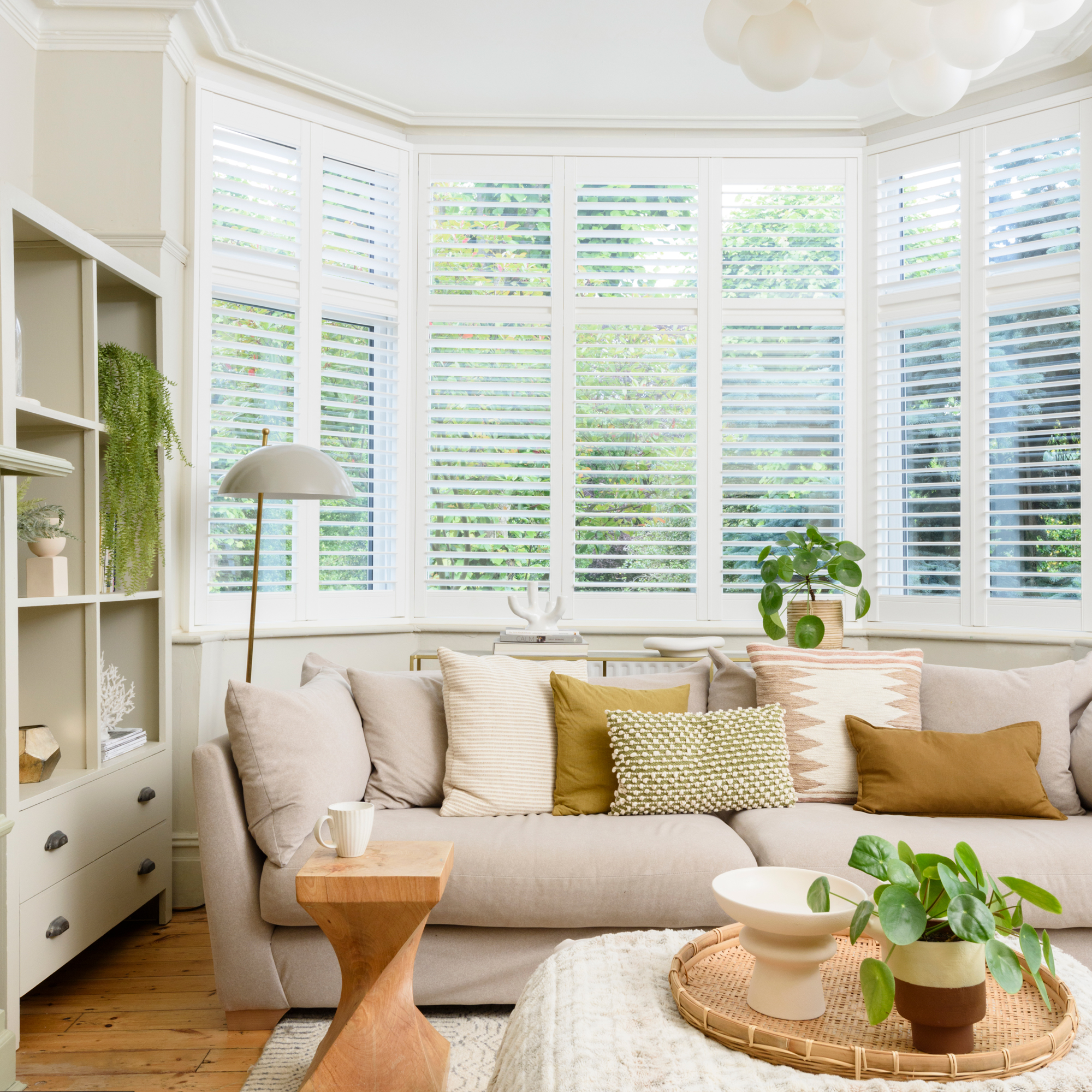 a neutral coloured living room with a large three seater beige sofa covered in scatter cushions, beside a bay window with fitted shutters