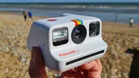 En hand som håller upp en Polaroid Go-kamera på en strand.