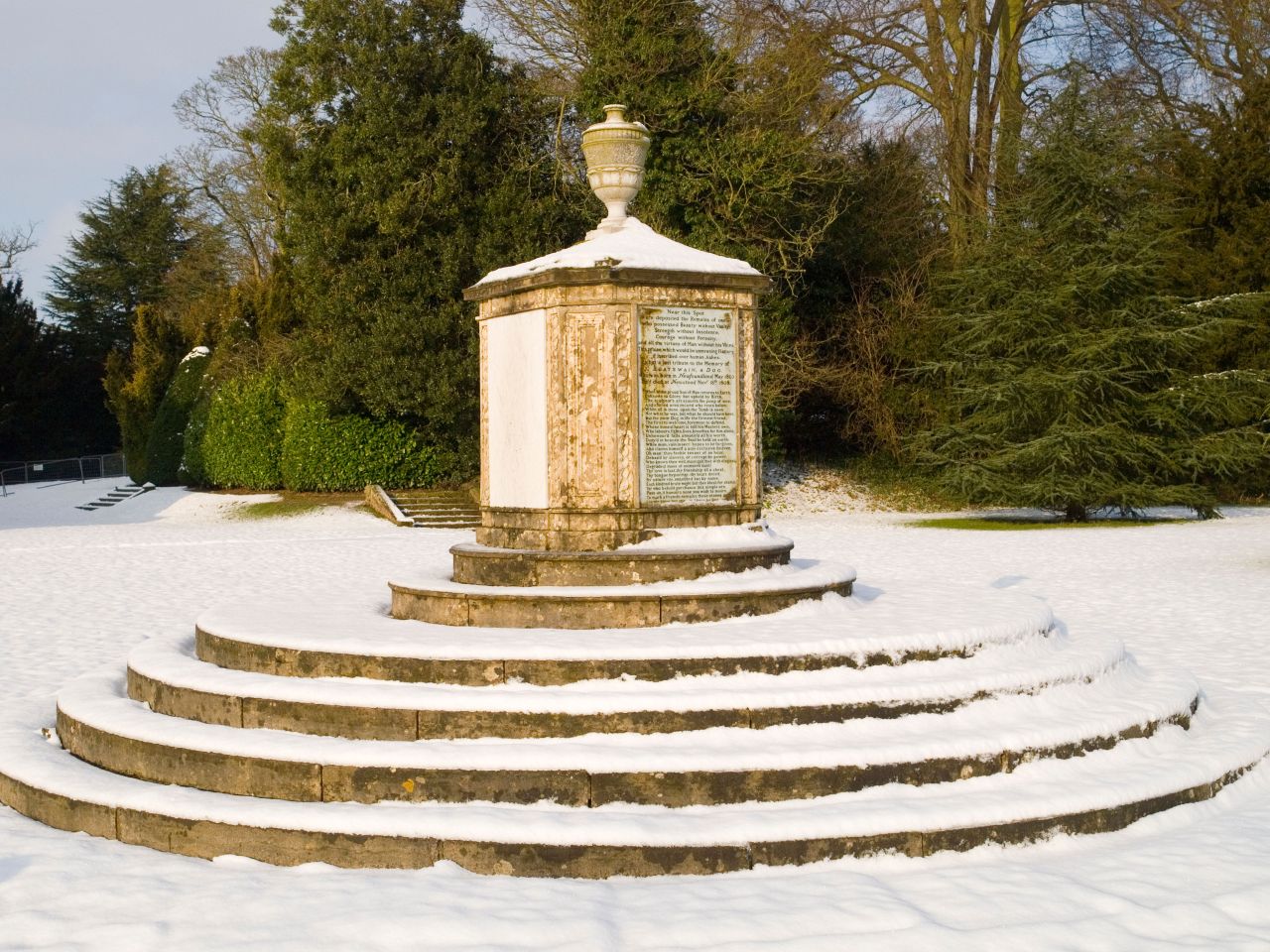 A stone monument set on circular steps stands in a snow-covered garden surrounded by trees.