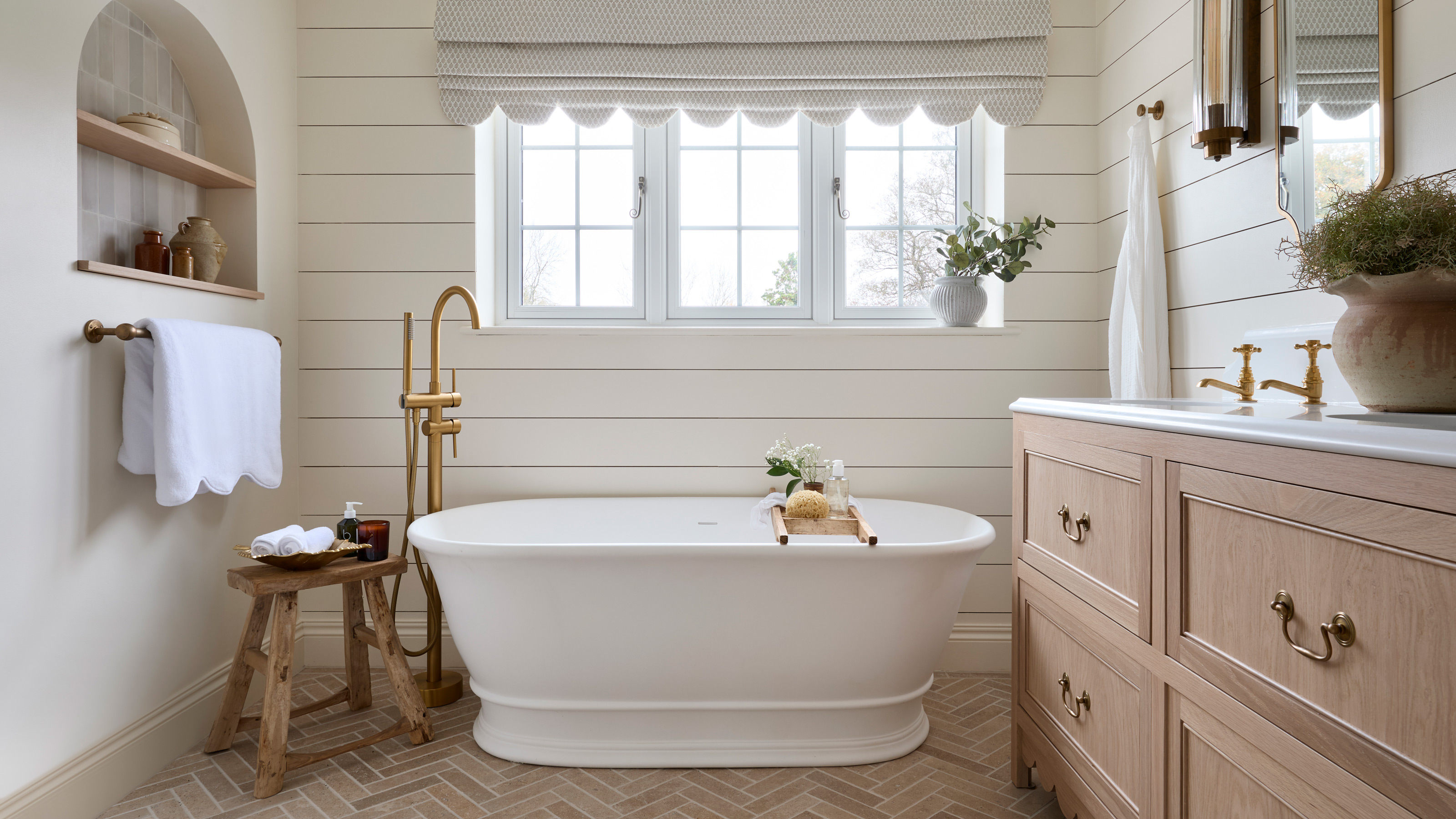 a neutral bathroom with horizontal wall panelling, a large white freestanding bath tub and brass fixtures, double vanity unit, a scalloped edge roman blind and limestone herringbone floor tiles