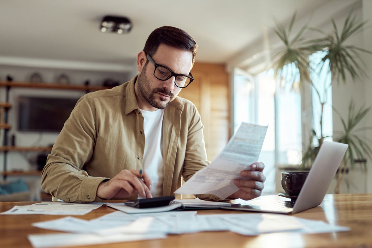 Man holds financial document as he uses calculator at desk beside laptop.