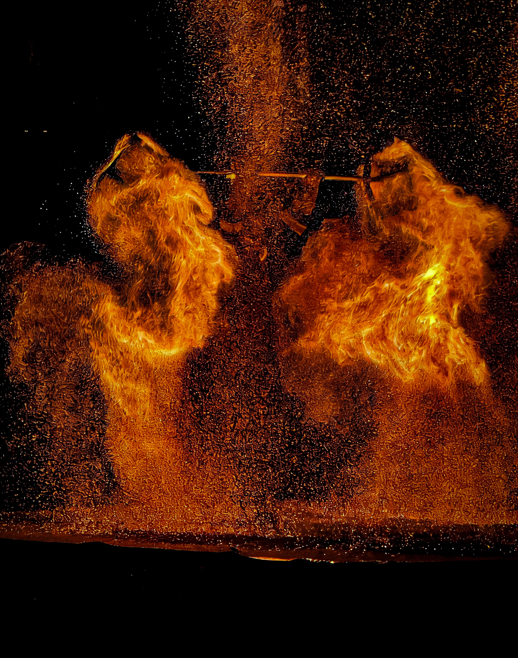 A night scene of a dramatic performance with molten iron or fire being thrown into the air, creating glowing orange explosions of sparks and flames shaped like wings against a black background