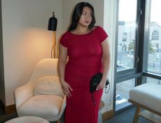 Woman standing in hotel room in a red, short-sleeve maxi dress and black clutch bag.