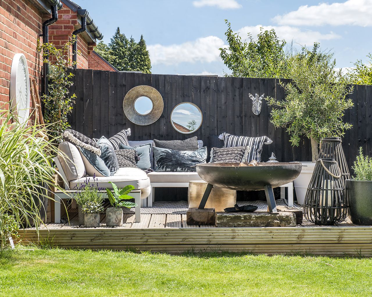 A decked patio area in front of a black fence with attached mirrors and grey sofa