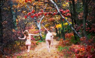 Two naked women running down a path in the woods, surrounded by autumn foliage