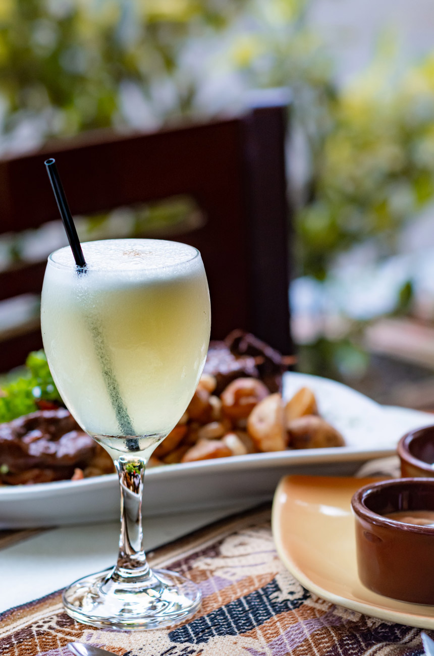 A cocktail on a table with a plate of food and a chair in the background