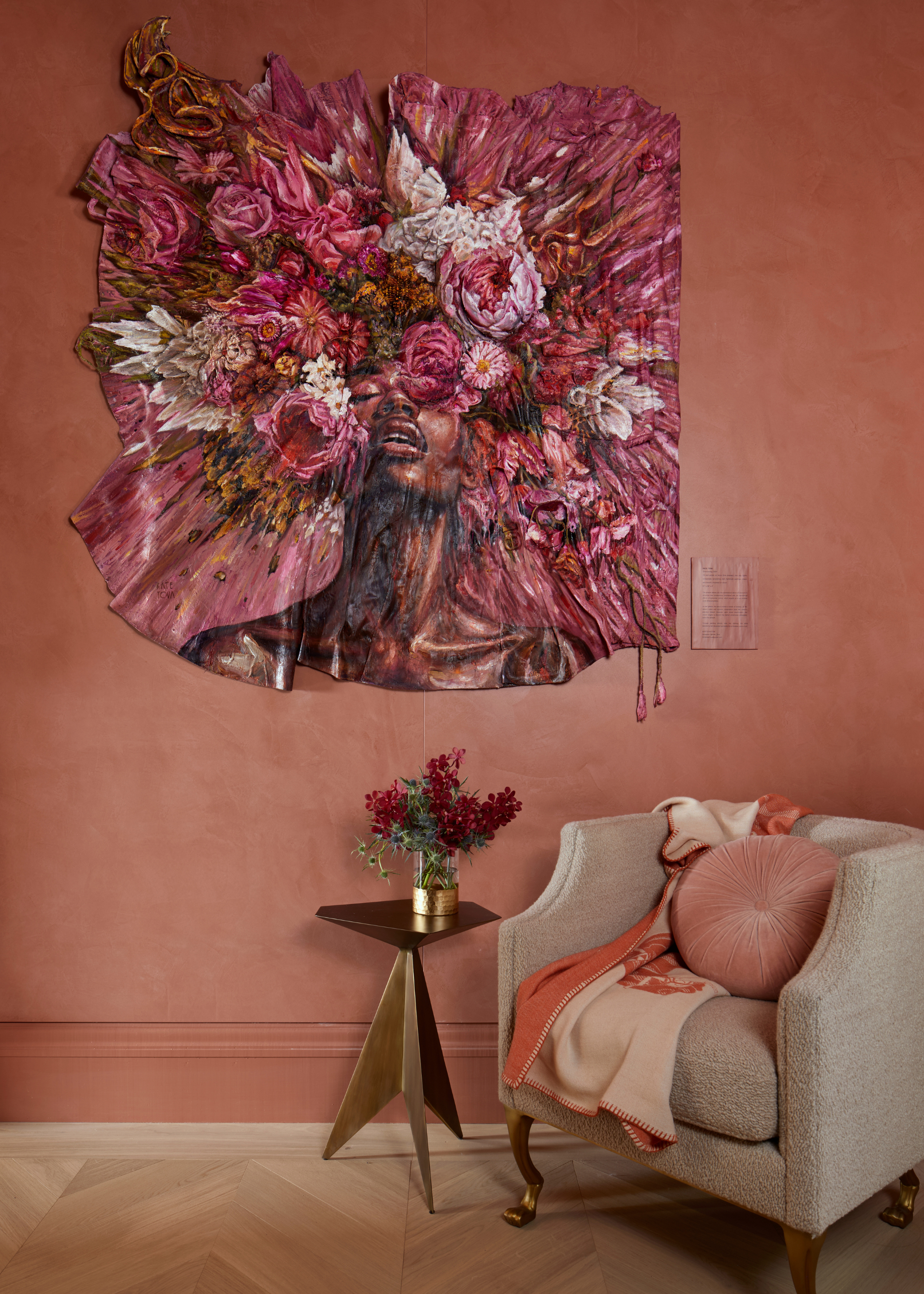 A dusty pink dressing room with a sofa seater, a plush pillow and a two-toned scarf, alongside a side table with a vase of flowers