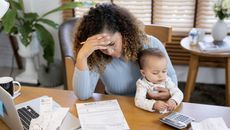 A young mother looks overwhelmed as she holds her baby and looks over financial paperwork at the dining room table.