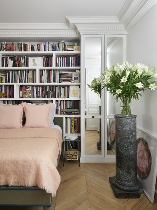 small apartment bedroom with mirrored doors, plinth with flowers, bookcase behind bed, herringbone wood floor