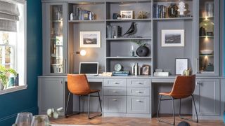 A dual home office with grey shaker cabinetry, two desks, leather chairs, open shelving and warm lighting, set against rich teal walls and parquet flooring.